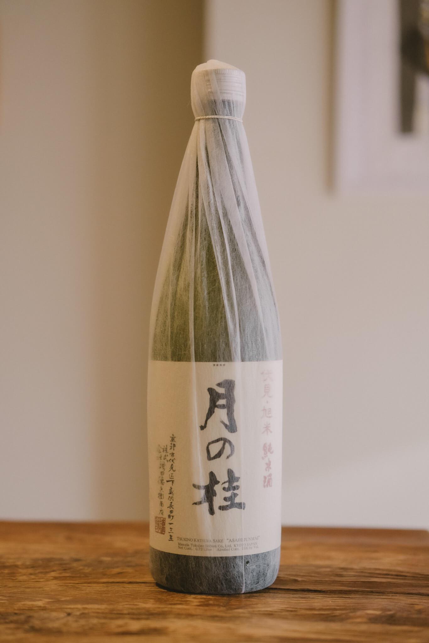 warm image of japanese wine covered in what looks like cream rice paper, bottle placed on dark oak wood table, with cream wall as background. Asian letters written in black on the front of the bottles label.