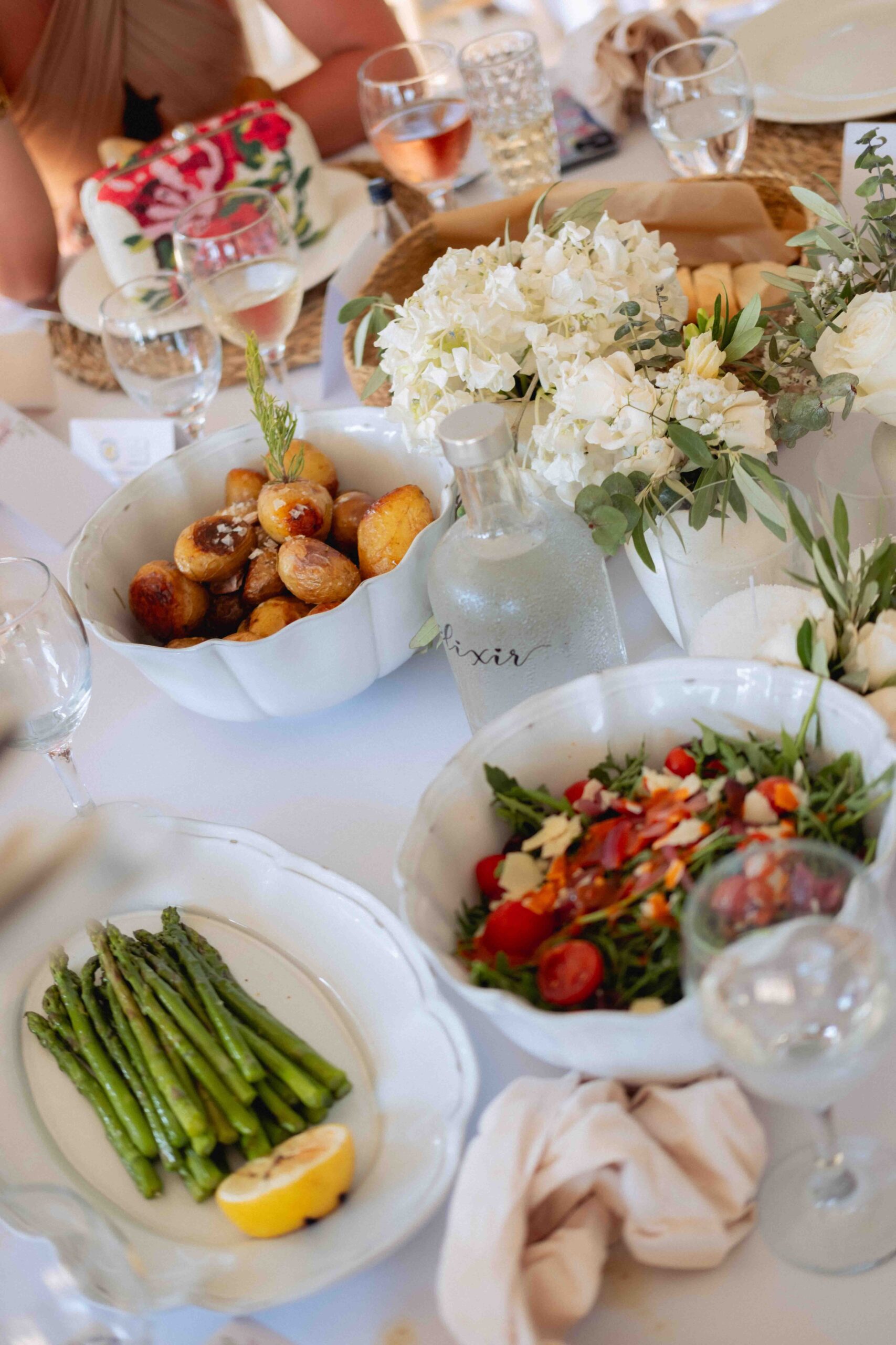 close up food photography of wedding table set up. Plate of vibrant green asparagus, tomato salad and roasted potatoes at ibiza elixir wedding. clear bottle of water and cream flowers also decorating the table.