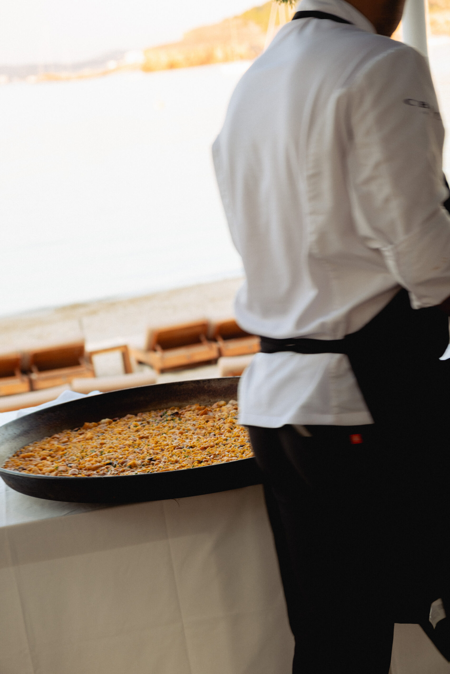 abstract, interesting photograph of paella chef standing next to a large paella dish with views of the ocean at CBBC in ibiza. hes wearing a white shirt and black trousers with a black apron. 