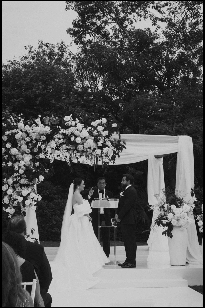 Bordered | Documentary Wedding Photographer Mimi Lambley-4 black and white film looking image taken with fujifilm in play del carmen, Mexico. white American couple standing at the alter getting married destination wedding. Big flower arrangements decorating the alter with white mesh fabrics