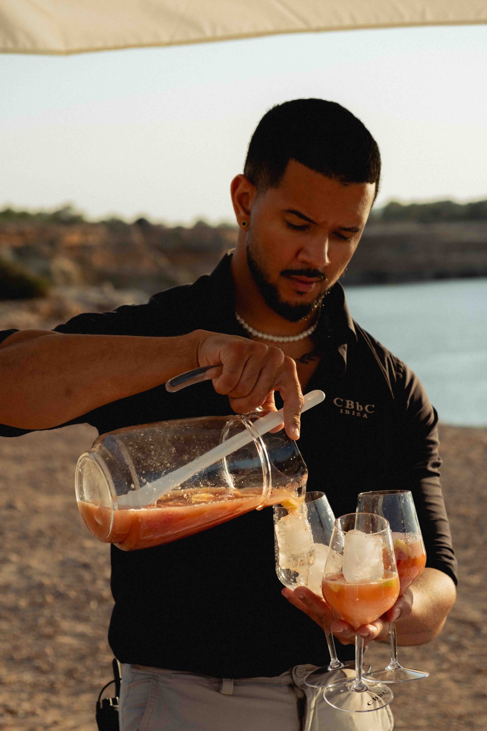 photograph of a black man pouring sangria from a litre jug into three wine glasses at sunset, ocean and ibiza cliffs in the background, blue skies and warm tones.