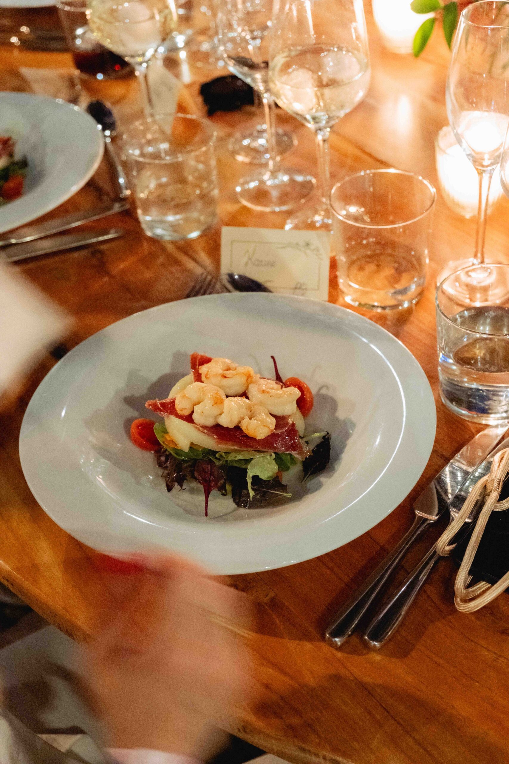 warm photograph of a plate of food on a table, blurry hands in motion in frame, glasses and cutlery scattered around the dining table at CBBC wedding dinner 
