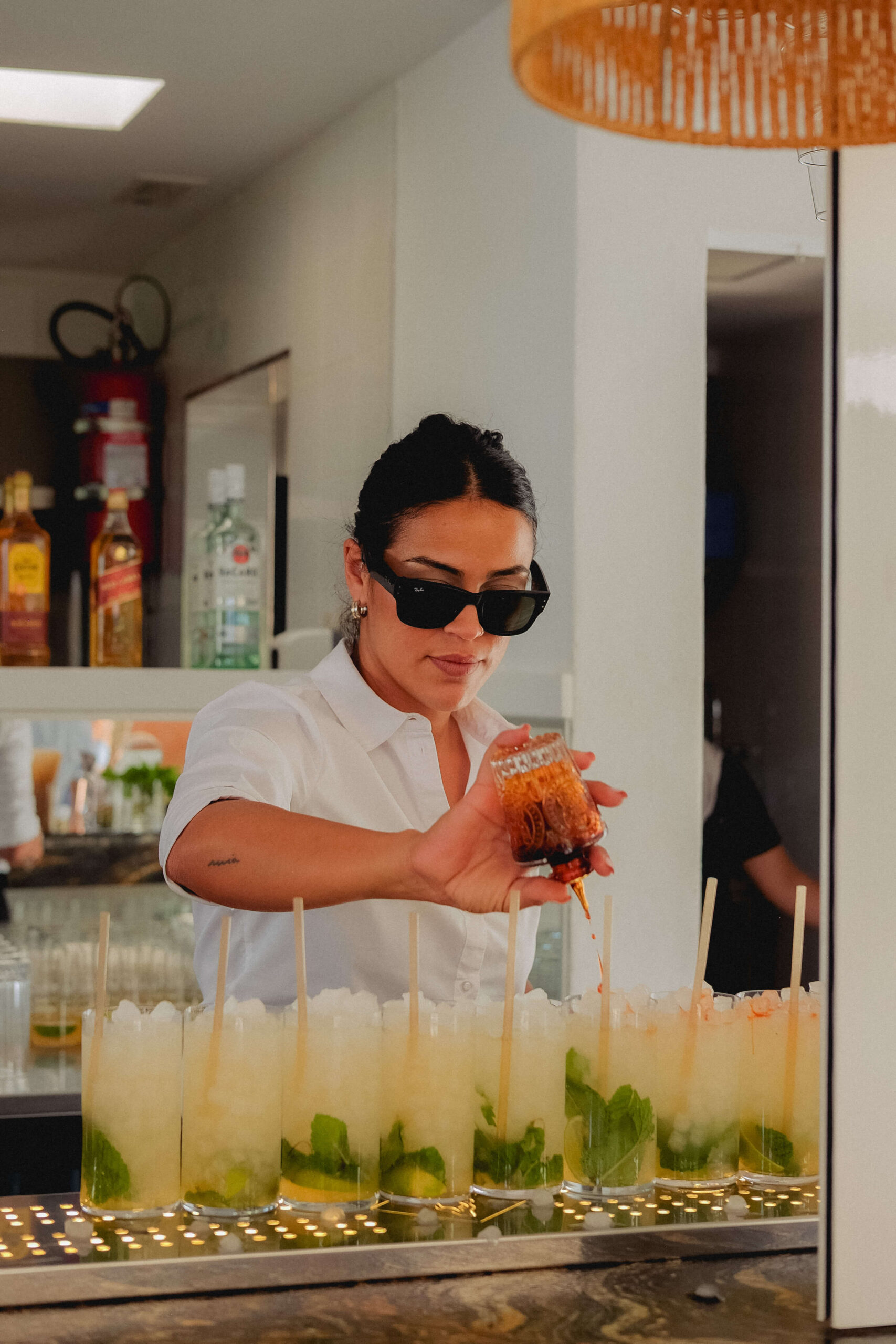 latina woman pouring and making eight cocktails at the bar at wedding event. she wears white shirt, slick back dark black hair and black sunglasses on with a straight face in ibiza